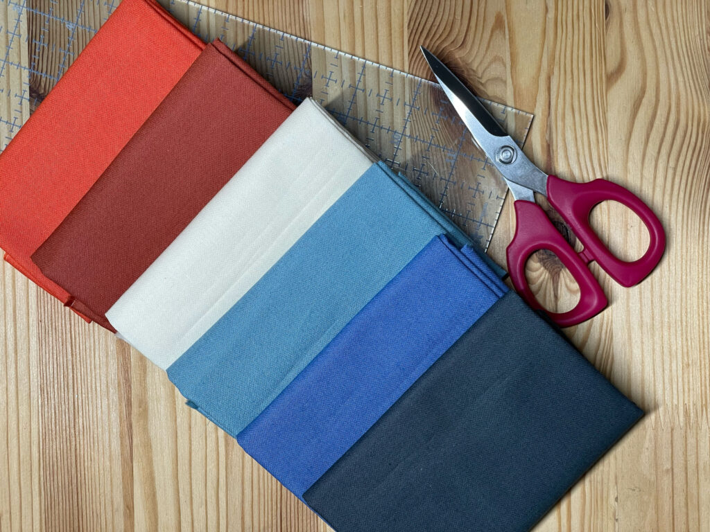 Fat Quarters in red, rust, light blue, cornflower and navy sitting on a ruler next to scissors